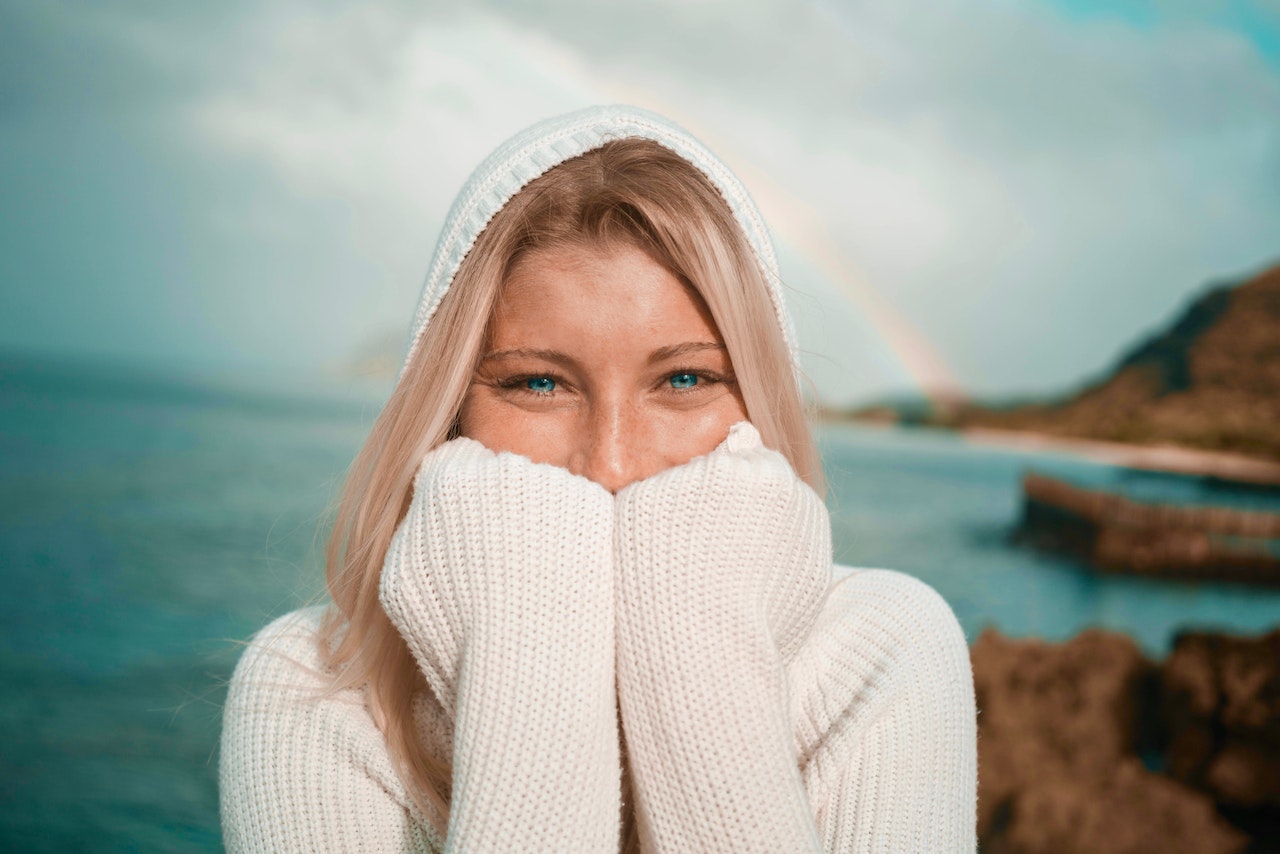 A happy woman in a  woolen sweater amidst the backdrop of a rainbow over a blue green sea and rocky coastline