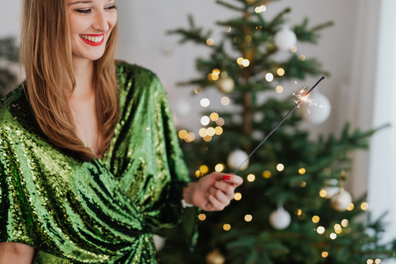 A happy and healthy woman smiling in a green shiny top while holding a sparkler near a Christmas tree