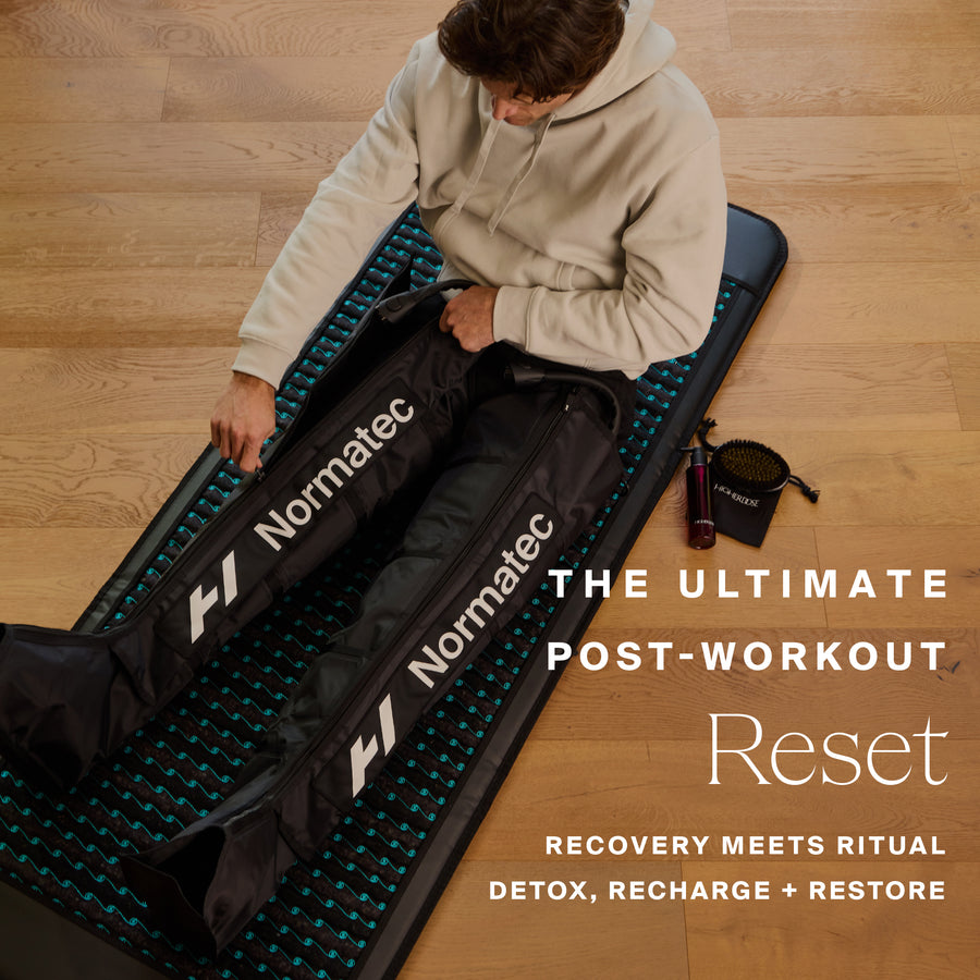 Person seated on blue and black Normatec recovery mat with compression leg wraps and recovery product bottle on wooden floor.