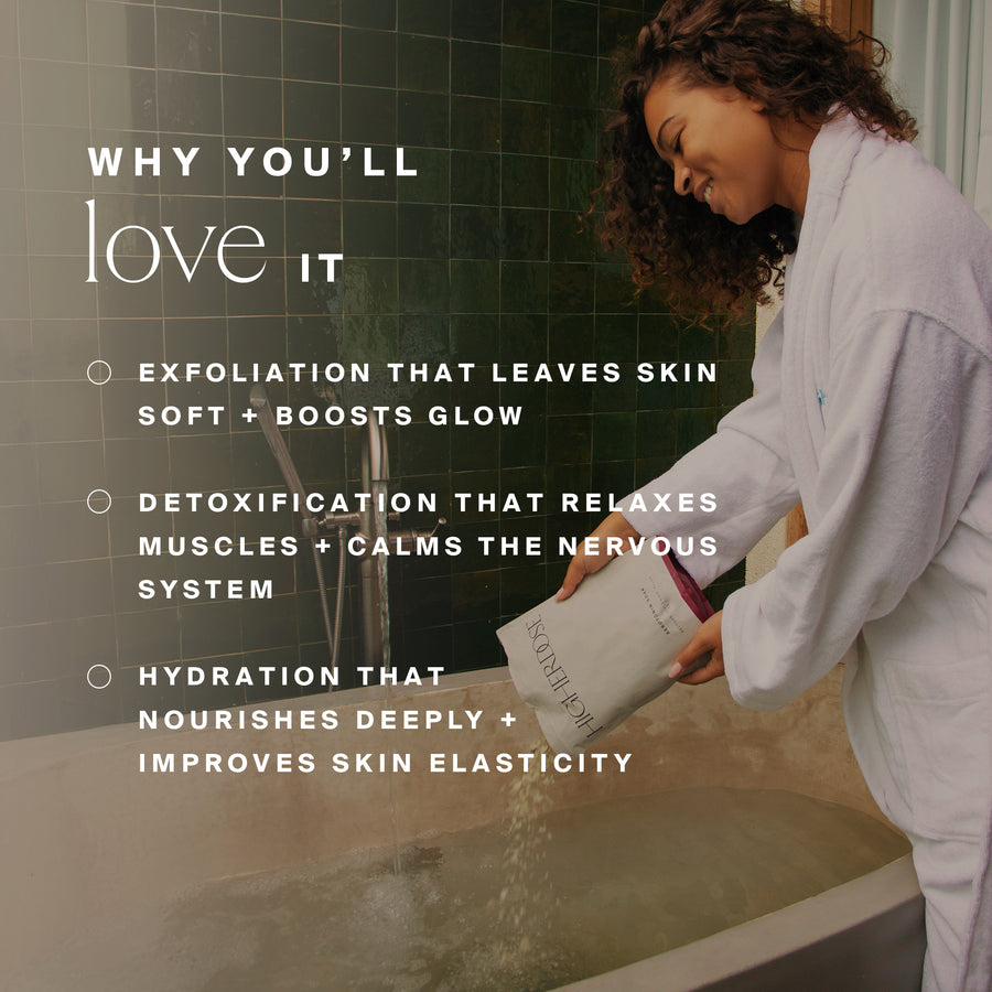 Woman in lavender shirt holding skincare product box while standing at bathroom sink with green tile wall.