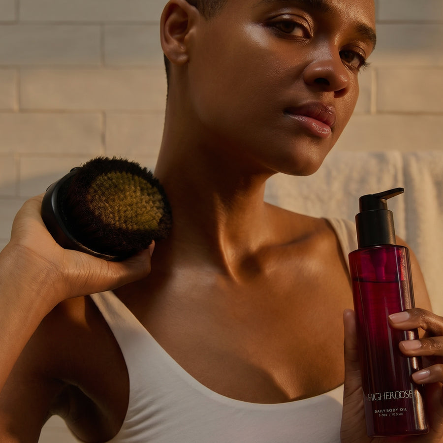 Woman holding a black circular body brush and a burgundy pump bottle labeled 