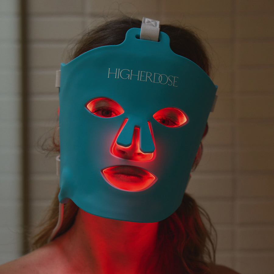 Teal silicone face mask with red LED light therapy elements on eyes, nose, and mouth areas, worn by person with dark hair.