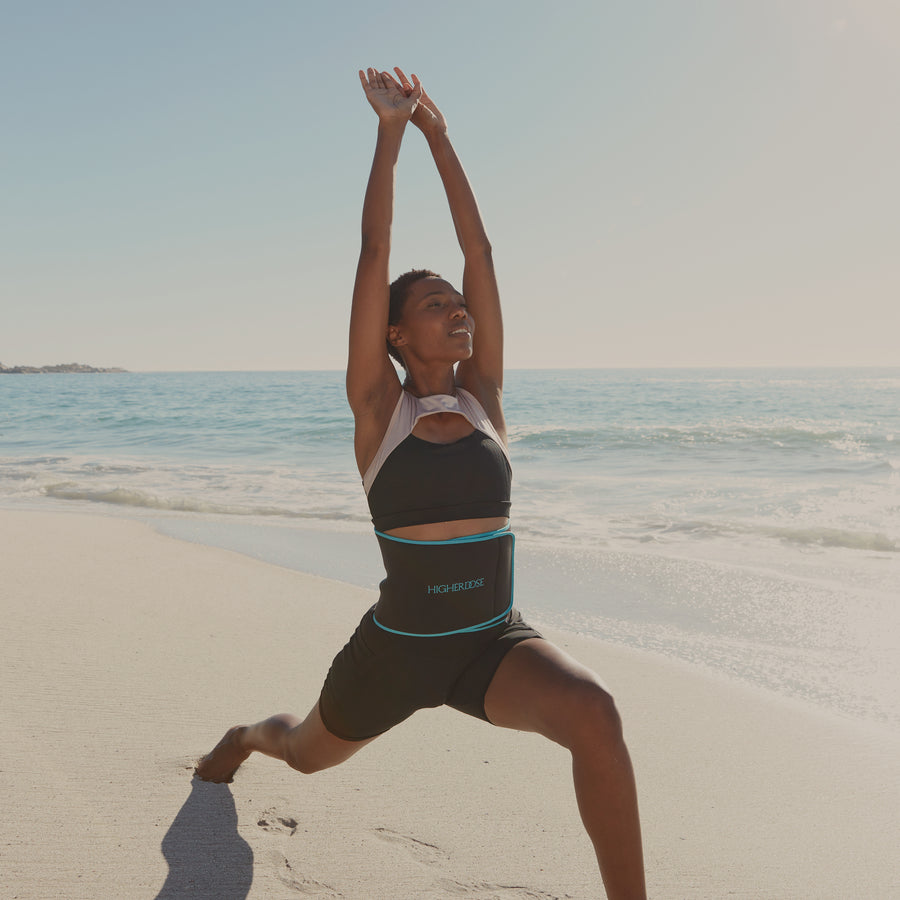 Woman wearing black sports bra and shorts with teal-trimmed waist band performing yoga pose on beach with ocean in background