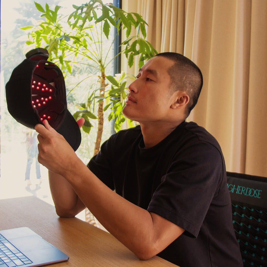 Man holding black device with red LED lights beside potted plant at desk.