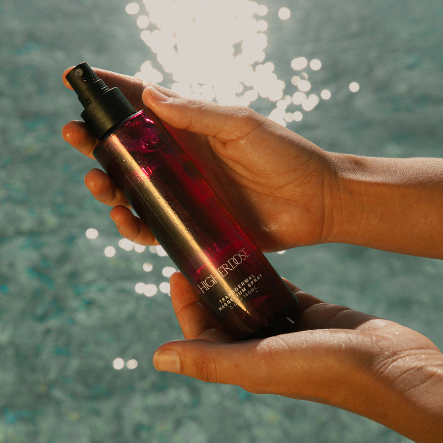 Dark glass spray bottle with gold and burgundy labeling held in two hands against a blurred turquoise water background.
