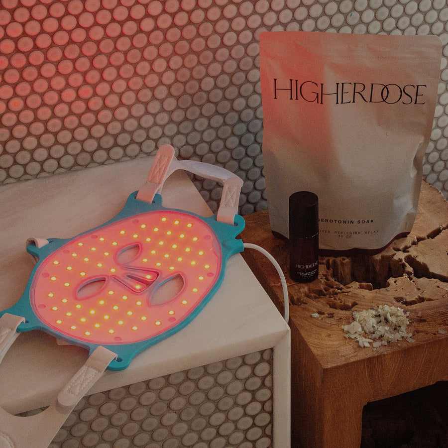 Pink and yellow silicone face mask on white cloth next to Higher Dose product pouch and dark bottle on wooden surface.