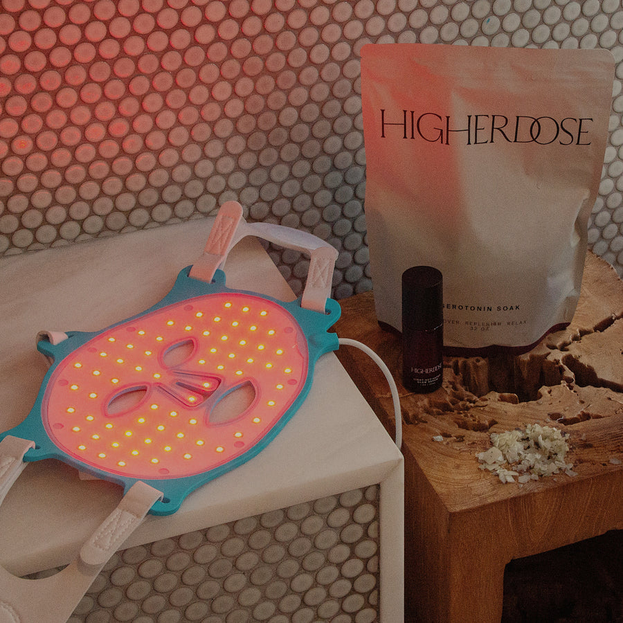 Pink and yellow silicone face mask on white cloth next to Higher Dose product pouch and dark bottle on wooden surface.