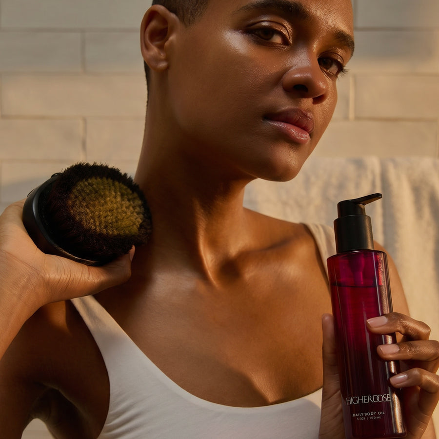 Person holding a black textured brush and red liquid product bottle with pump dispenser against neutral background.