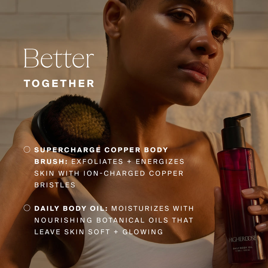 Person holding a copper body brush and dark red body oil bottle against neutral bathroom background.