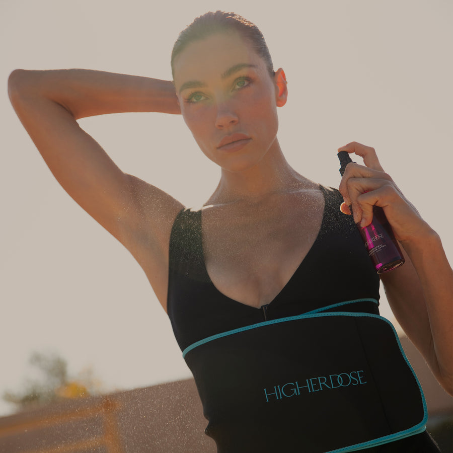 Woman wearing black sports top with turquoise trim holding purple spray bottle, with visible perspiration on skin and body.