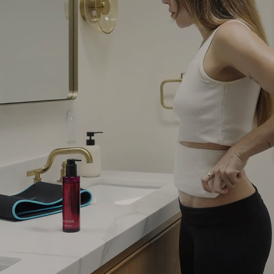 Red pump bottle, black resistance band, and white container displayed on bathroom counter next to person in white crop top.