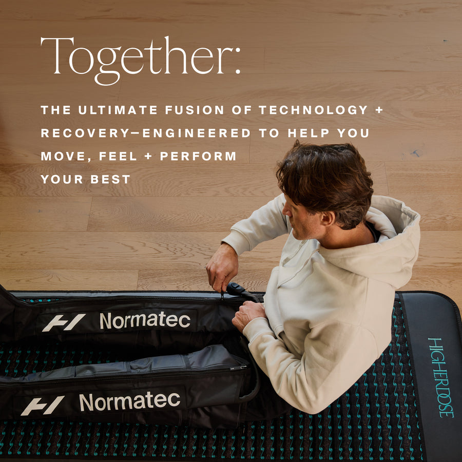 Person in cream hoodie using two black Normatec recovery leg sleeves on wooden floor with teal mat.