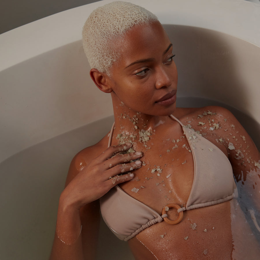 Woman in white bikini top sitting in bathtub with sea salt or exfoliating scrub on shoulders and chest.