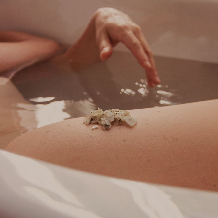 Hand applying granular exfoliating scrub to arm skin over a bowl of water.