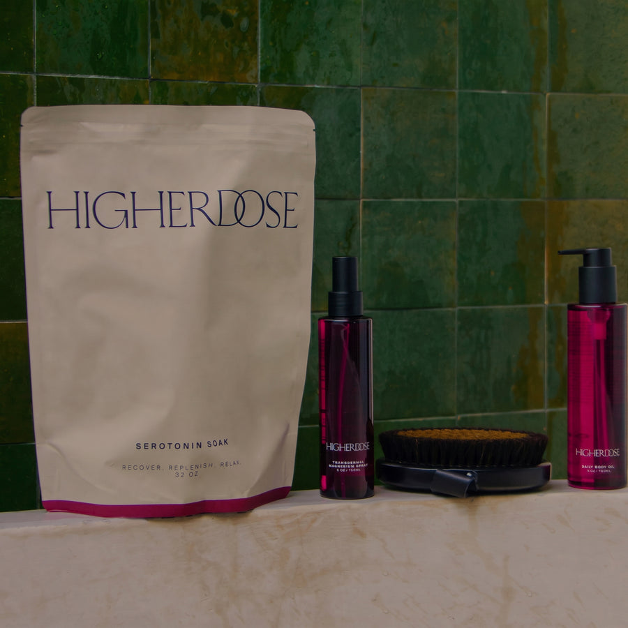 Higher Dose product set with cream pouch, two red spray bottles, and dark circular brush on green tile background.