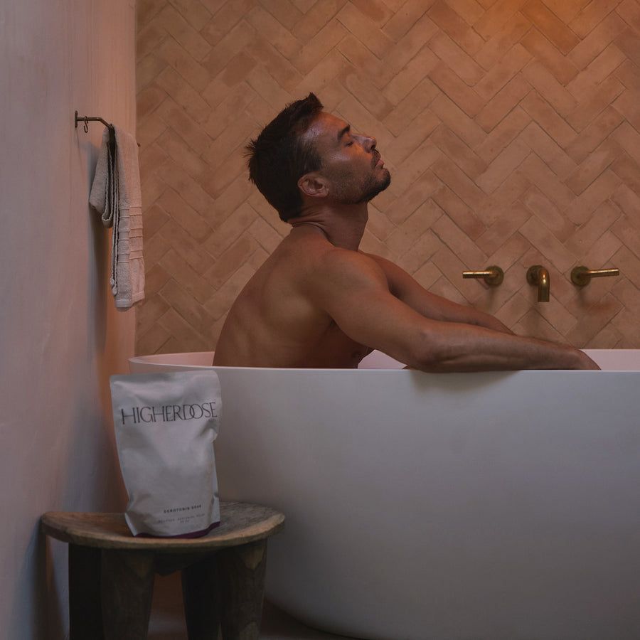 Man relaxing in white bathtub with HIGHERDOSE branded pouch on wooden stool beside tub.