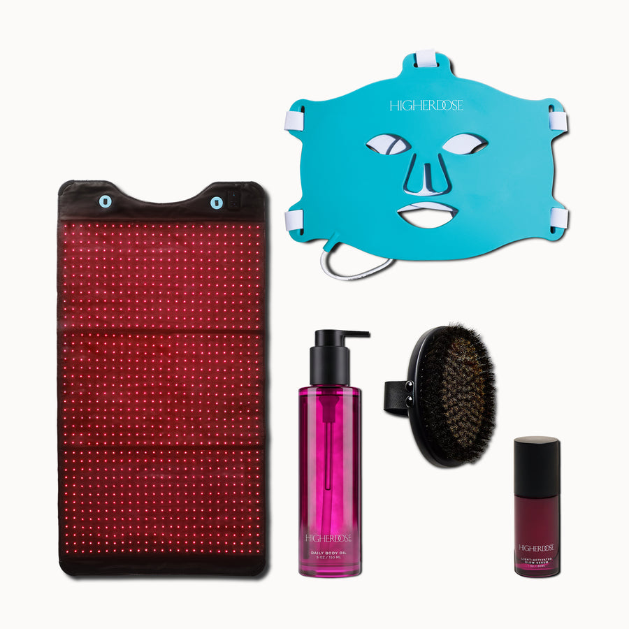 Skincare set with red light therapy panel, turquoise face mask, magenta body oil bottle, black brush applicator, and burgundy