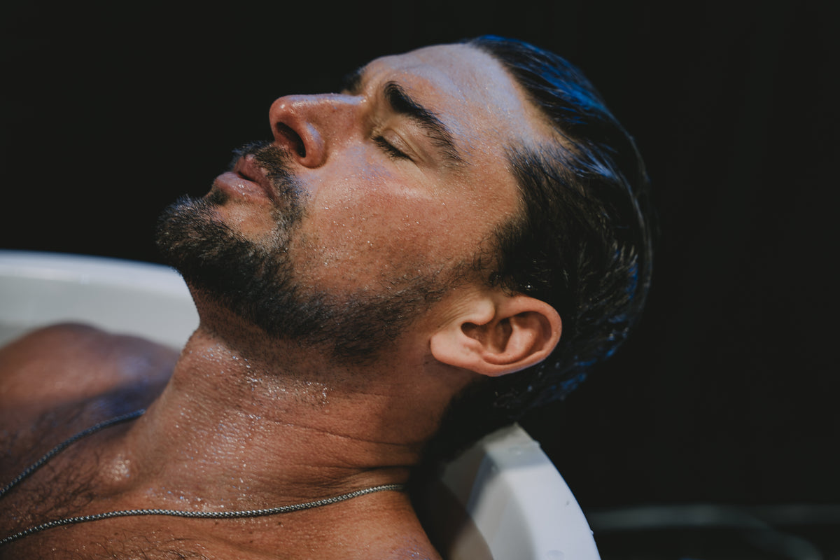 Man with a beard and wet hair lying on a white surface against a dark background