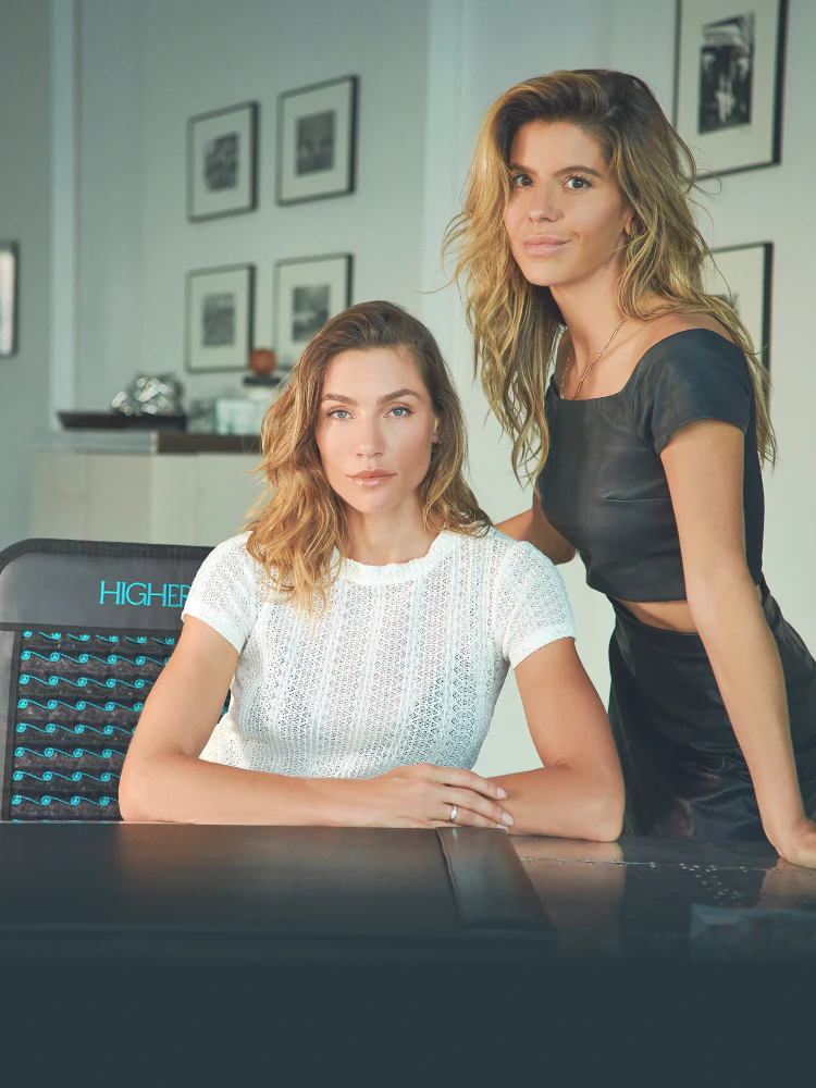 HigherDOSE founders Lauren Berlingeri and Katie Kaps pose together indoors beside a HigherDOSE infrared mat.