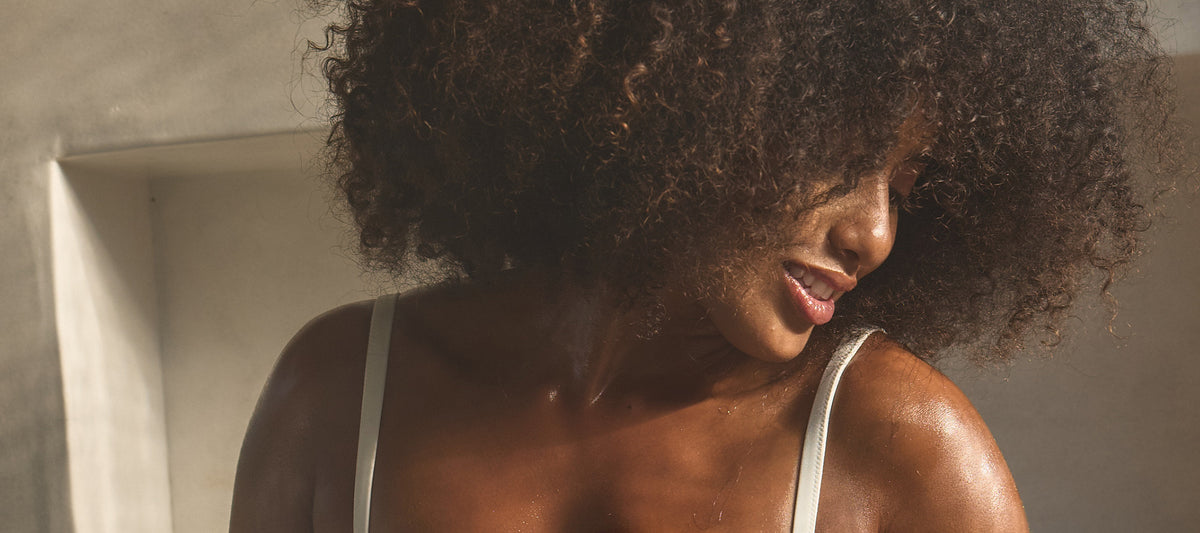 A woman with voluminous curly hair smiles softly while looking downward, her skin glistening with moisture in warm, natural light.