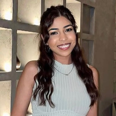 Woman smiling indoors, wearing a sleeveless light blue top.