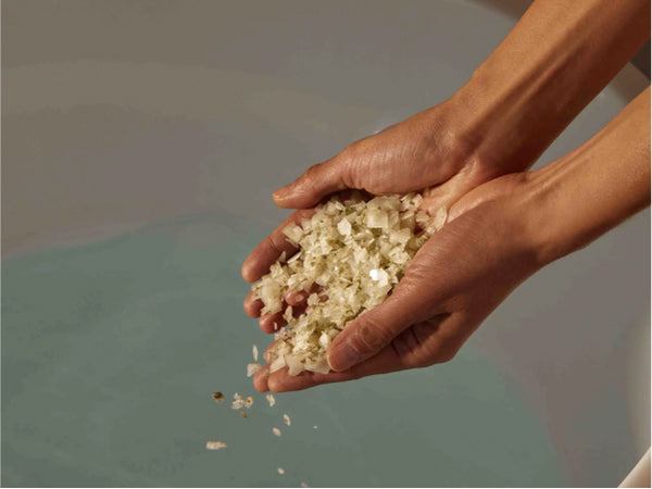 Hands holding a scoop of bath salts above a tub filled with water.