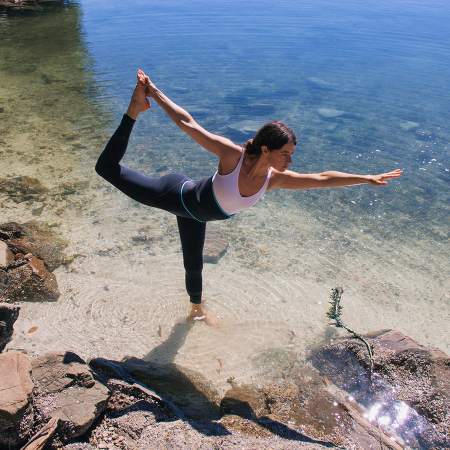 Woman in white tank top and black leggings performing yoga pose in shallow water at beach.
