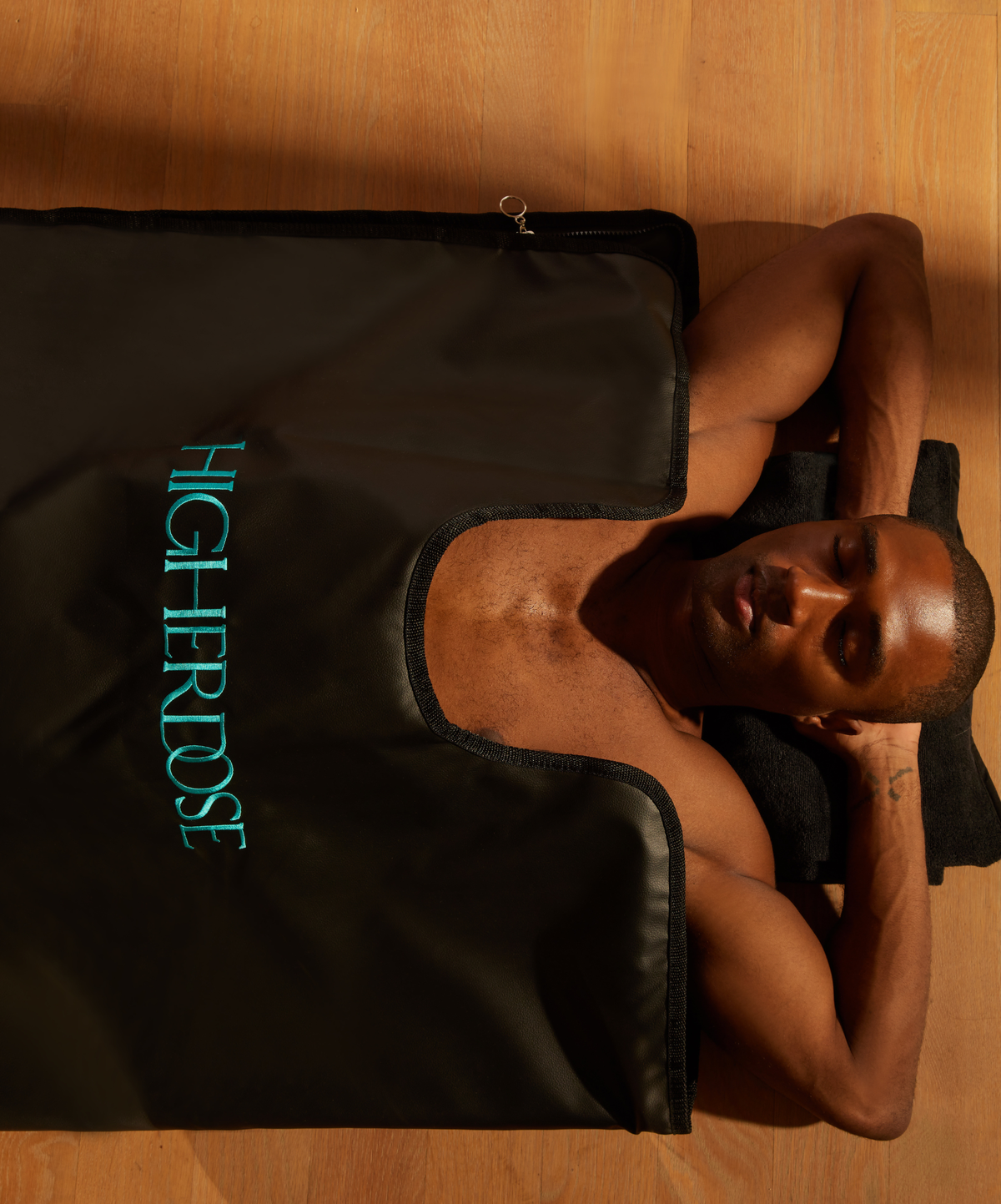 A man lies on a black heated panel pad on a wooden floor, wearing a black tank top with the brand name 'HIGHHEROSE' printed in teal on the chest.