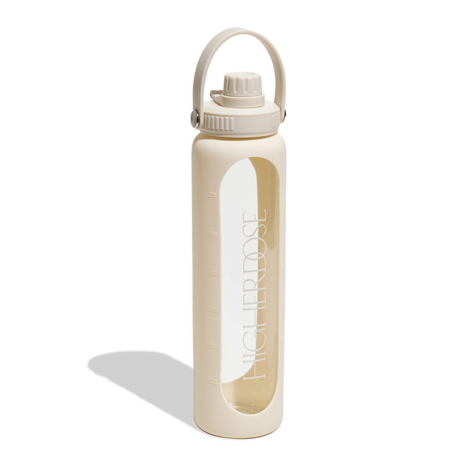 Cream-colored glass water bottle with beige silicone sleeve, carrying handle, screw-top lid, and time markers on the glass in