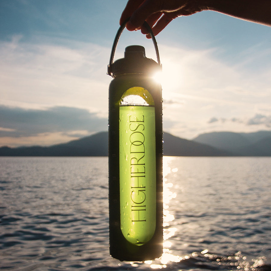 Hand holding a HigherDOSE green water bottle against the sunlight over a lake.