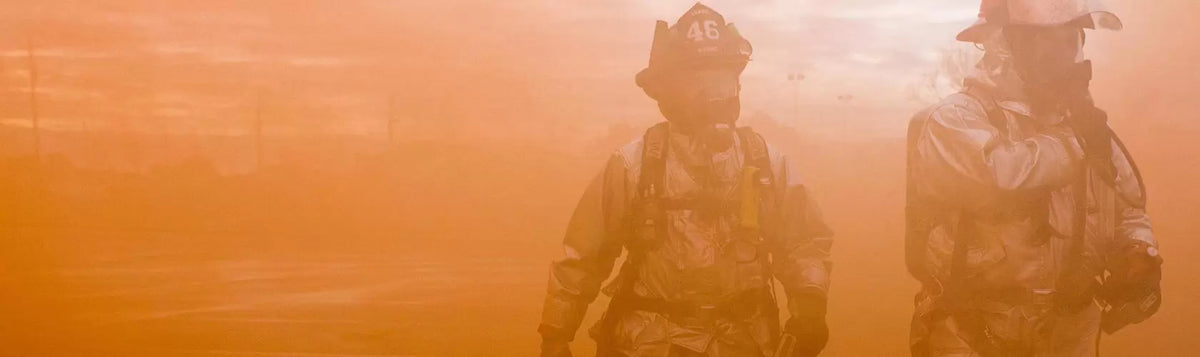 Two firefighters in gear walk through thick orange smoke, partially obscured by haze and light.