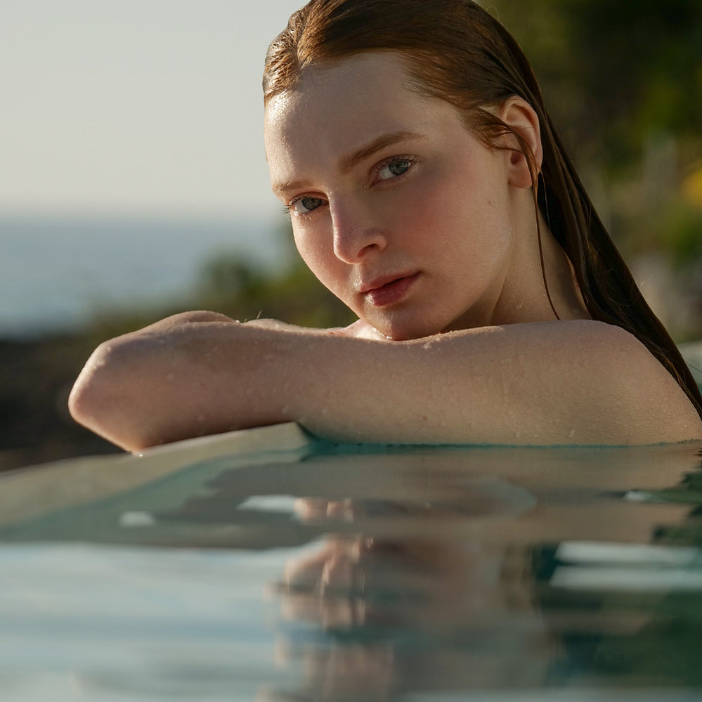 Woman relaxing in a pool, leaning on the edge and gazing toward the camera, embodying the HigherDOSE lifestyle.